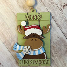 Load image into Gallery viewer, Layered wooden Merry Christmoose Christmas ornament featuring a brown moose wearing a red Santa hat and blue striped scarf against a green background.
